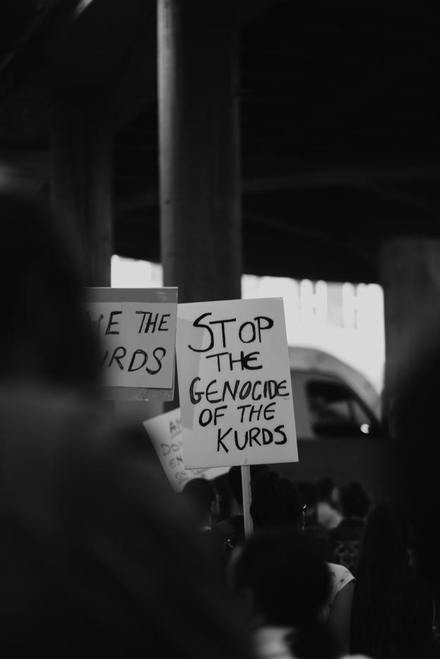 Eine Gruppe von Menschen steht vor einem Gebäude und hält Schilder mit der Aufschrift "Stoppt den Völkermord an den Kurden" in einem Schwarz-Weiß-Bild, mit einem Fahrzeug im Hintergrund.