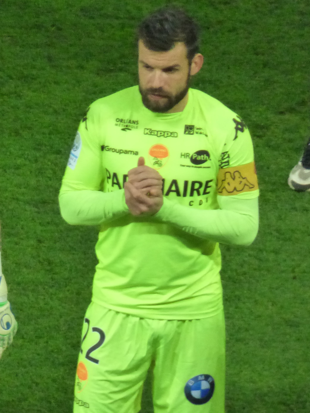 Ein Mann in einem gelben Torwarttrikot, der ein grünes T-Shirt und Shorts trägt, steht auf einem Fußballfeld und klatscht in die Hände, wobei auf der linken Seite des Bildes ein weiteres Bein zu sehen ist.