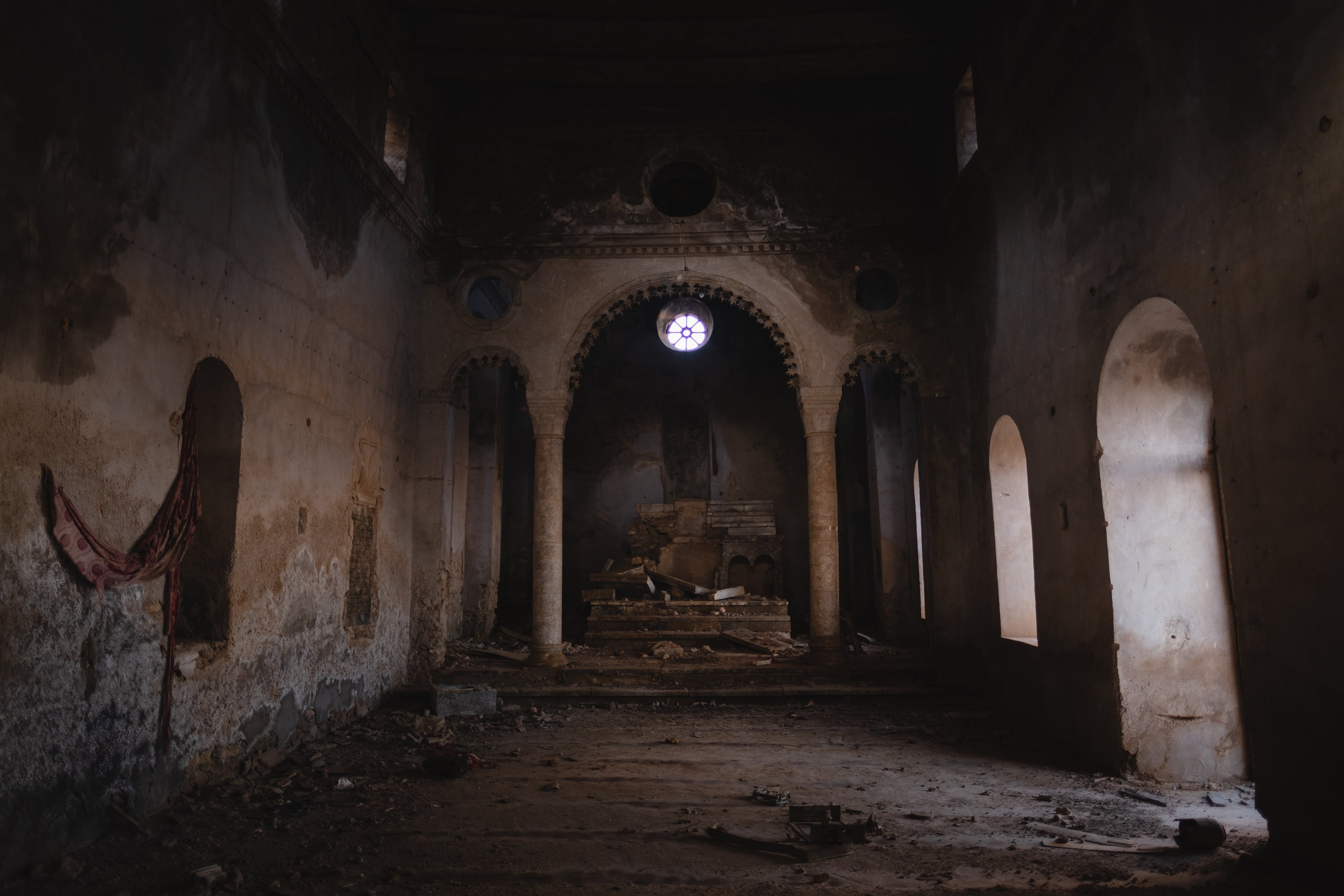 Alter Bau mit Bögen, Säulen und einem mittigen Fenster, mit einem drapierten Tuch an der linken Wand und verstreuten Gegenständen auf dem Boden, der eine verlassene Kirche mit dunkler, unheimlicher Atmosphäre vermittelt.