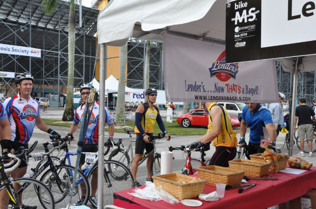 Eine Straßenszene mit Menschen in der Nähe von Fahrrädern, ein Geschäft mit Gegenständen auf einem Tisch, Bäume, Zelte, an einem Schuppen angebrachte Banner, Fahrzeuge und Gras.