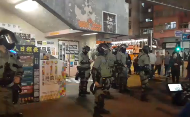 Eine Gruppe von Polizisten in Uniform steht vor einer Menge von Demonstranten mit Helmen und Waffen, mit Plakaten an den Wänden, Lampen an der Decke und Gebäuden mit Fenstern im Hintergrund, in Hong Kong.