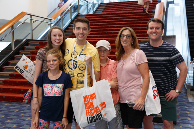 Eine Gruppe strahlender Menschen mit Taschen steht zusammen vor einer Rolltreppe auf der Nationalen Buchmesse 2019, mit Treppen, Geländern und einer Wand im Hintergrund.