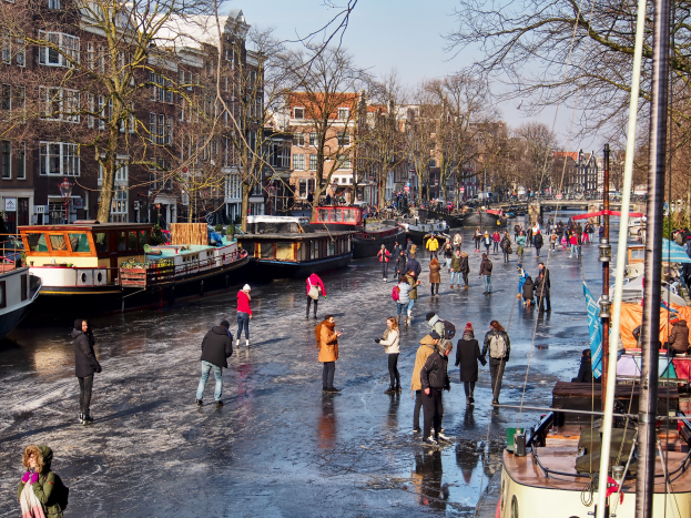 Eine Gruppe von Menschen, die auf einem Kanal in Amsterdam Eislaufen gehen, umgeben von Booten, Bäumen, Gebäuden mit Fenstern, Laternenmasten und einem bewölkten Himmel.