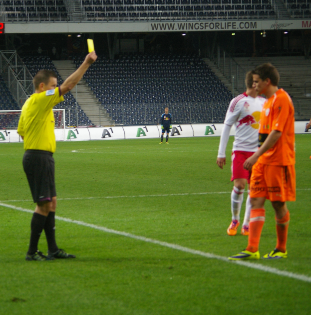 Ein Fußballschiedsrichter zeigt einem Spieler auf dem Feld eine gelbe Karte, umgeben von einer Gruppe von Menschen und einem Torpfosten, einem Zaun, einem Schild, Metallrahmen, einem Anzeigebildschirm, Stühlen und einer Decke mit Deckenleuchten im Hintergrund.