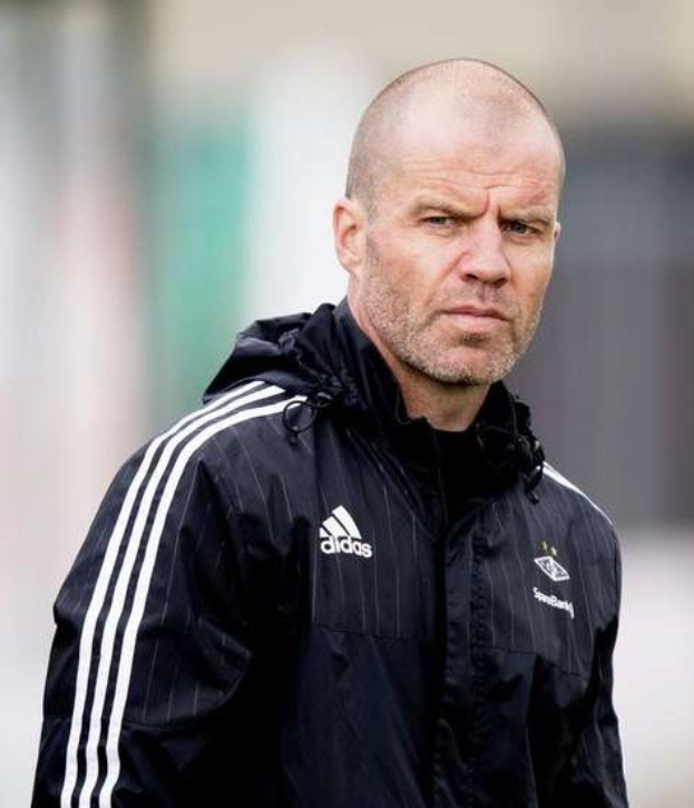 Ein kahlköpfiger Mann in einer schwarzen Jacke steht auf einem Fußballfeld, trägt eine marineblaue Jacke mit weißen Streifen und hat die Hände verschränkt.