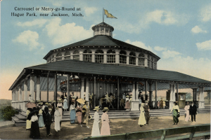Eine alte Postkarte zeigt ein Karussell im Hague Park, Jackson, Michigan, mit Menschen, einige halten Schirme, ein Gebäude mit Säulen und einer Flagge, Bäume und einen bewölkten Himmel, mit Text oben.