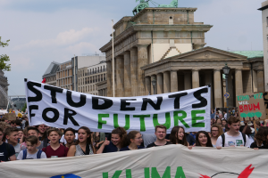 Eine Gruppe von Studenten marschiert in Berlin, eine bunt bemalte Fahne schwingend, auf der "Students for Future" steht, mit Gebäuden, Bäumen und Himmel im Hintergrund.