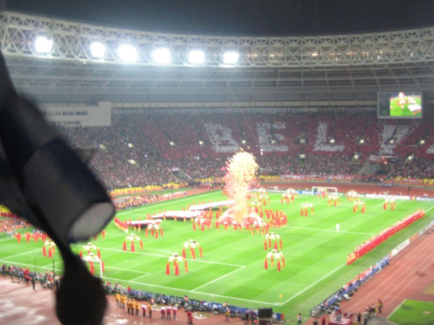 Ein Stadion mit Flutlicht, Eisengittern, Zuschauern, stehenden Personen, Werbetafeln, Anzeigetafeln, Überwachungskameras und diversen Dekorationen einschließlich Flaggen.