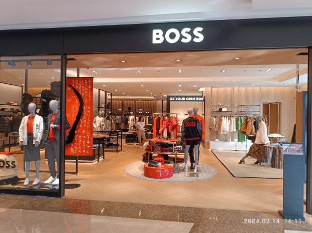 Das Innere eines Hugo Boss Stores in einem Einkaufszentrum mit Schaufensterpuppen in Hugo Boss Kleidung, Kleider an Bügeln, Tischen mit verschiedenen Gegenständen, beleuchteter Decke und einem Textschild oben.