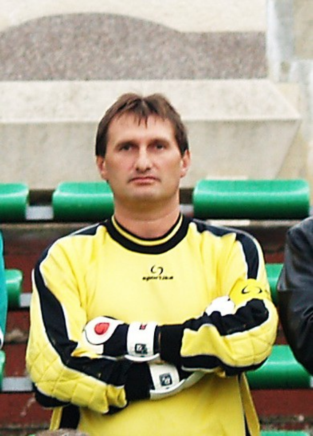 Zwei Männer stehen sich gegenüber; der Mann in einem gelben T-Shirt, der wie ein Torwart aussieht, hat einen entschlossenen Gesichtsausdruck, während der andere einen schwarzen Blazer trägt. Grüne Stühle und eine Wand sind im Hintergrund zu sehen.