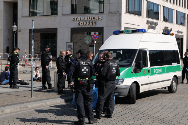 Eine Gruppe von Polizisten steht vor einem Starbucks-Café, rechts ist ein Van und links ein paar Menschen zu sehen; im Hintergrund sind ein Gebäude, ein Schild, ein Laternenmast und ein Zaun zu erkennen.