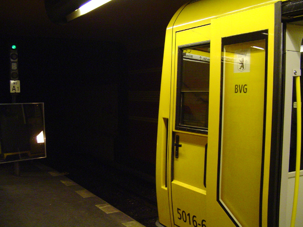 Ein Bahnhof mit einem Zug auf der rechten Seite, einem Pfahl und einer Tafel auf dem linken Bahnsteig, einer Lampe oben und Eisenbahnschienen unten.