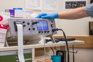 Eine Person in blauen Handschuhen arbeitet an einem medizinischen Gerät auf einem mit verschiedenen Gegenständen übersäten Tisch, mit Papieren an der Wand im Hintergrund, was auf eine Krankenhausumgebung hindeutet.