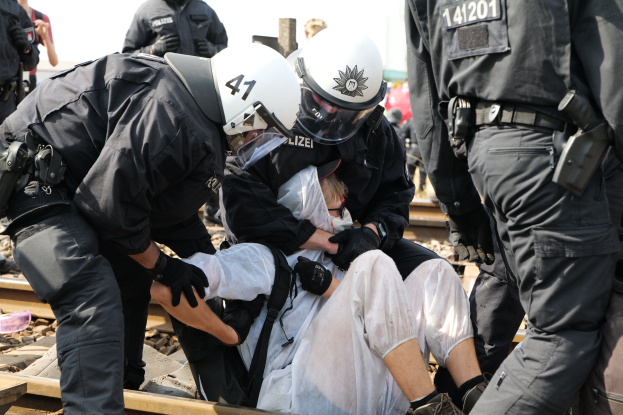 Eine Gruppe von Polizisten in schwarzer Einsatzbekleidung verhaftet einen Mann in einem weißen Kleid auf einem Bahngleis, mit anderen Menschen im Hintergrund.