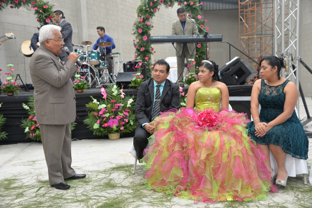 Ein Mann steht vor einem Mikrofon und spricht, während andere sitzen und ein Orchester im Hintergrund spielt, mit einer Bühne, die mit Blumen und Pflanzen geschmückt ist.