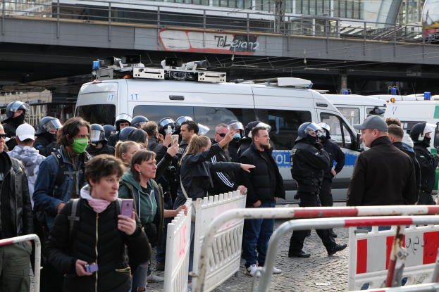 Gruppe von Menschen vor Polizeiwagen mit Barrikaden im Vordergrund, einige tragen Helme und halten Telefone, mit einer Brücke und Gebäuden im Hintergrund während einer Demonstration in Berlin, Deutschland.