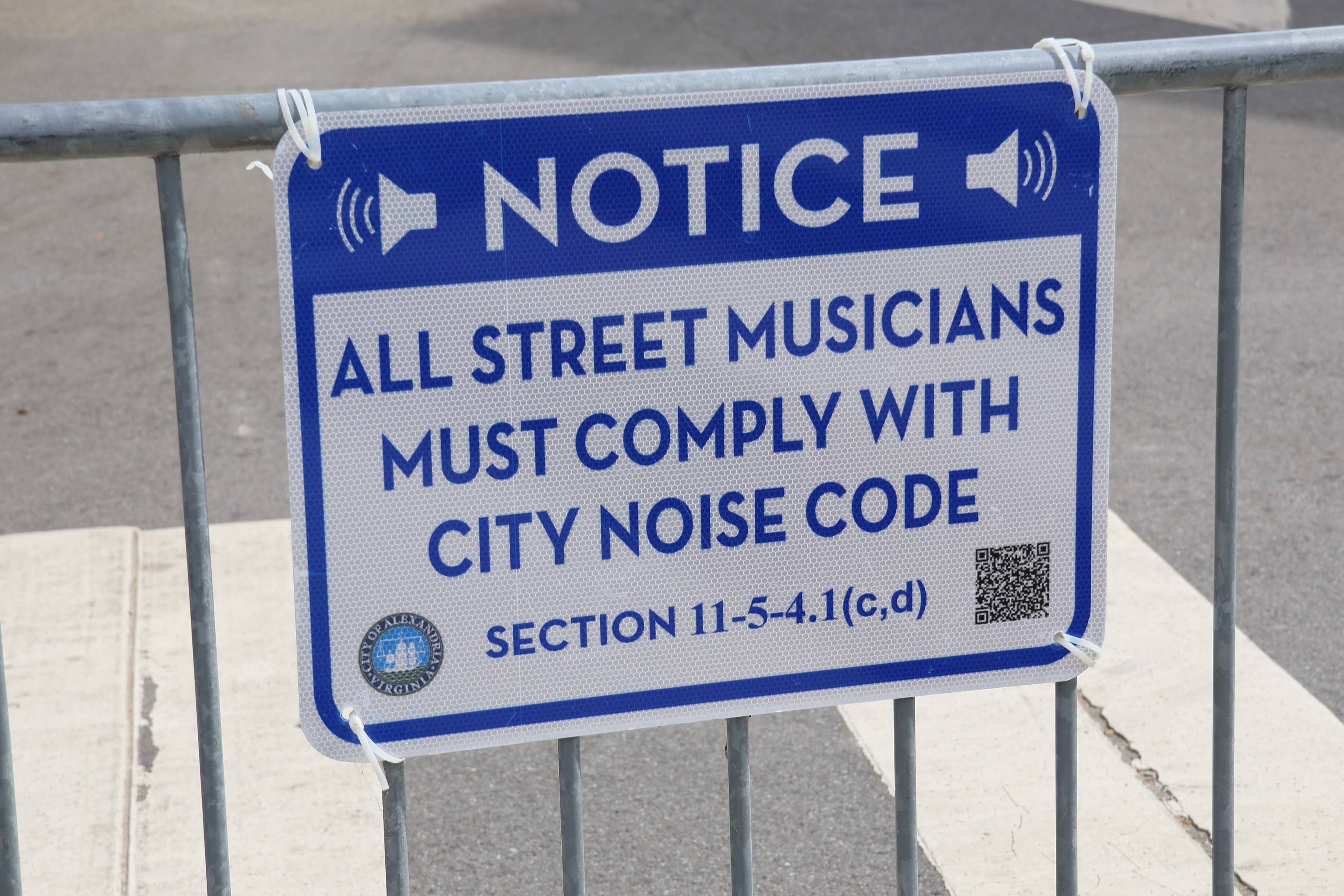 Anschlagtafel an einem Zaun mit einem Schild, auf dem "Hinweis: Alle Straßenmusiker müssen die städtische Lärmvorschrift einhalten" steht, und eine Straße im Hintergrund.