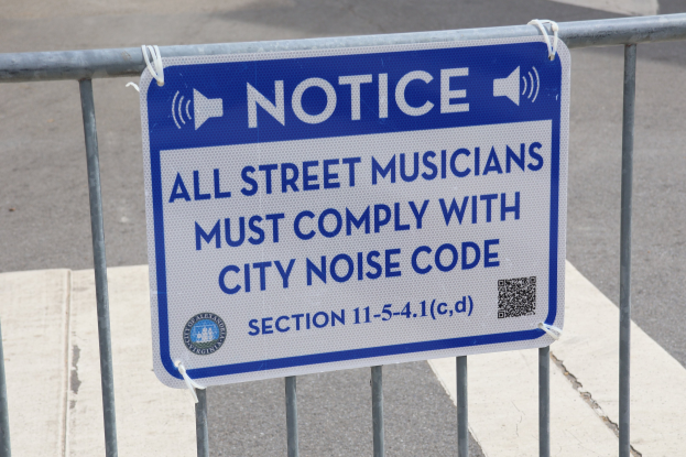Anschlagtafel an einem Zaun mit einem Schild, auf dem "Hinweis: Alle Straßenmusiker müssen die städtische Lärmvorschrift einhalten" steht, und eine Straße im Hintergrund.