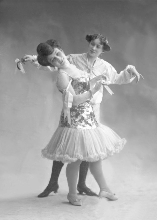 Zwei Frauen in einem Schwarz-Weiß-Foto tanzen zusammen, eine hält ein Band, gekleidet in abgestimmter Kleidung.