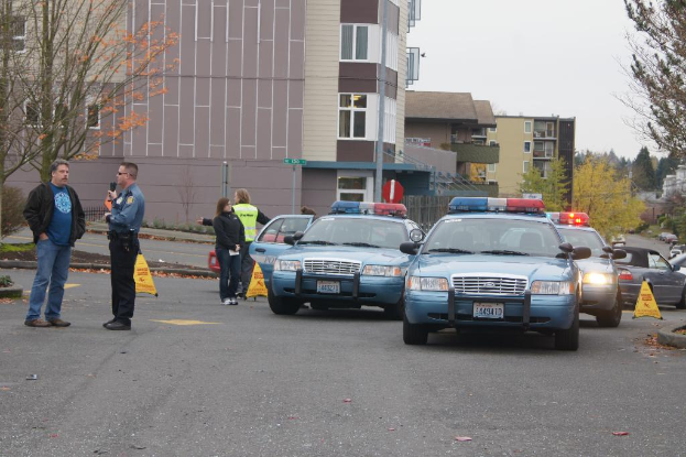 Autos auf einer Straße mit vier Menschen in der Nähe, Gebäude mit Fenstern im Hintergrund, Bäume und Warnblinkleuchten.