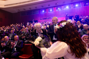 Eine Frau in einem weißen Kleid hält einen Blumenstrauß vor einer Menge sitzender, klatschender Menschen, mit einer beleuchteten Wand und einem Geländer im Hintergrund bei einer jüdischen Hochzeitszeremonie in London.