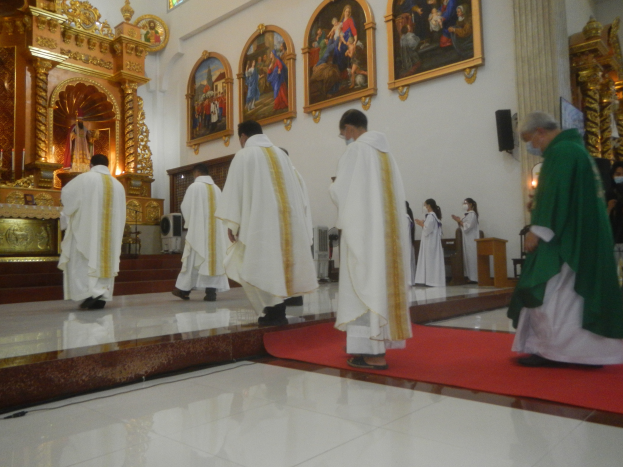 Eine Gruppe von Priestern geht einen roten Teppich in einer Kirche entlang, mit Menschen, die auf dem Boden vor ihnen und auf der rechten Seite stehen, und Foto Rahmen an der Wand im Hintergrund.