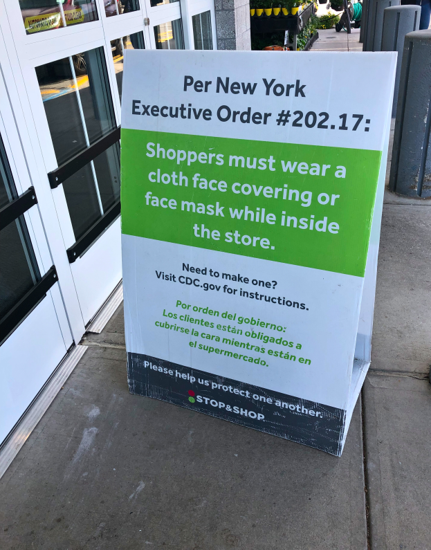 Ein Schild auf dem Gehweg vor einem Geschäft trägt die Aufschrift "Per New York Executive Order 202.17: Kunden müssen im Geschäft eine Stoffgesichtsbedeckung oder einen Mund-Nasen-Schutz tragen", mit einem Gebäude auf der linken Seite und Menschen, Pflanzen und Gegenständen auf der rechten Seite.