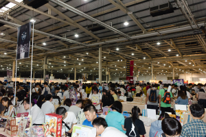 Eine große Gruppe von Menschen umringt Tische, die mit Büchern, Kisten und anderen Gegenständen gefüllt sind, auf einer Buchmesse, mit Bannern und Lichtern darüber.