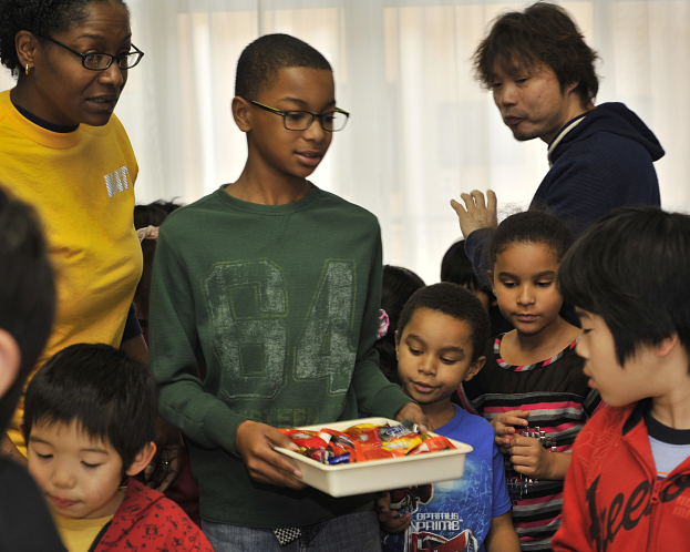 Eine Gruppe von Kindern steht zusammen in einem Raum mit einer Frau, die ein Tablett mit Schokoladen hält, und Vorhängen im Hintergrund.