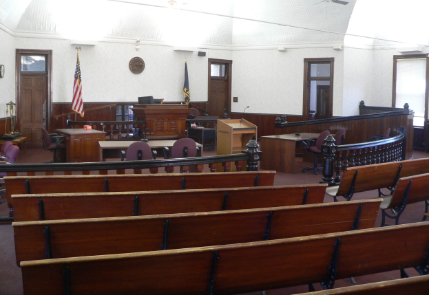 Ein Gerichtssaal mit hölzernen Kirchenbänken, Tischen, Stühlen, einem Podium, Fahnen, Türen, einer Uhr an der Wand und Lampen und Ventilatoren an der Decke.