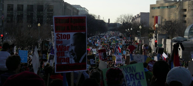 Eine Gruppe von Menschen, die auf einer Straße protestieren und Plakate halten, mit Gebäuden, Bäumen und Laternen im Hintergrund unter einem Himmel.