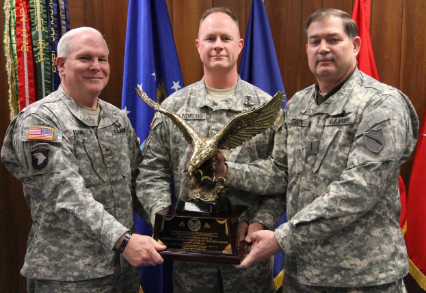 Drei Männer in Uniformen stehen mit Auszeichnungen, mit Flaggen dahinter und bunten Gegenständen mit Text oben links.
