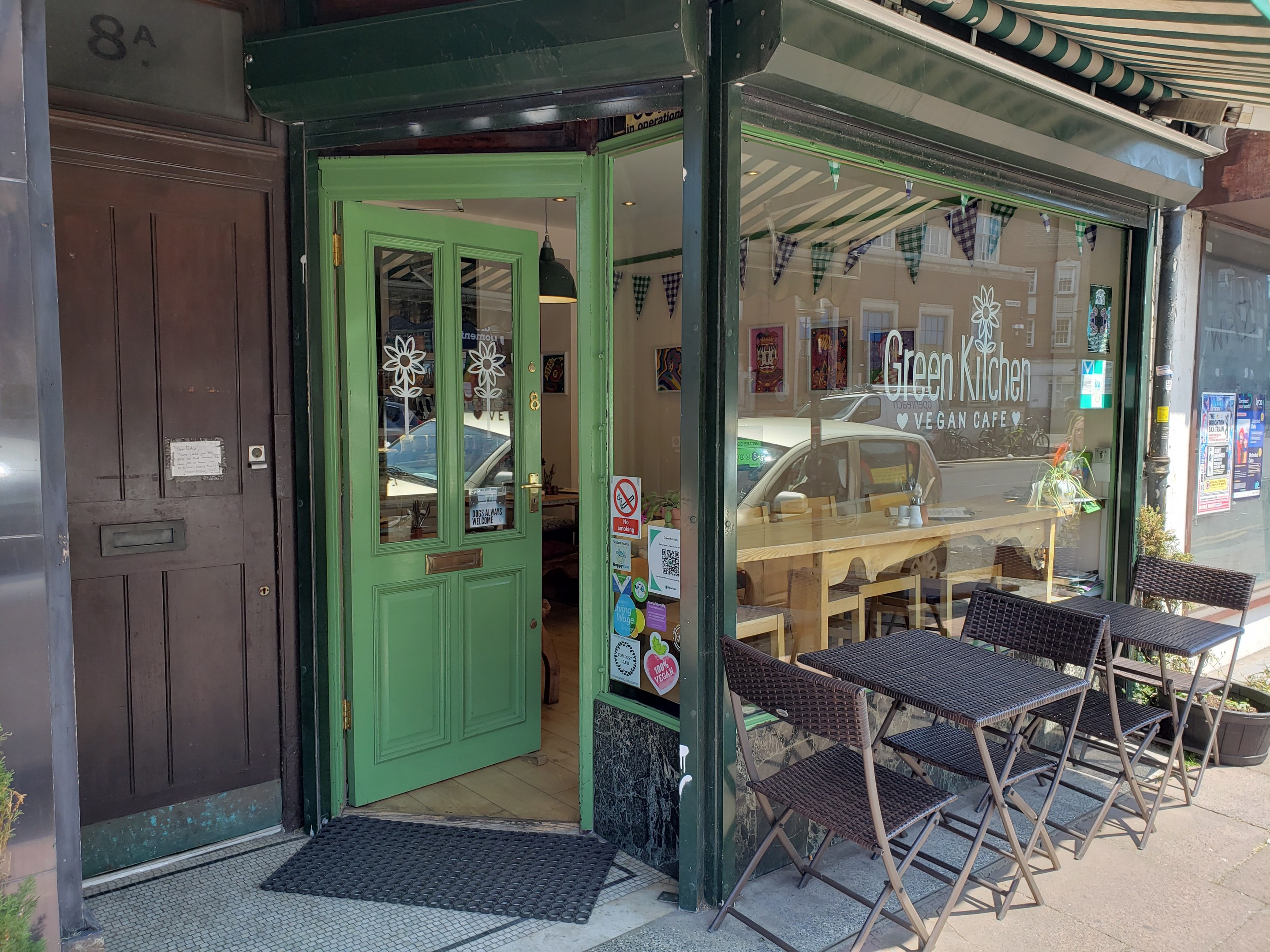 Außensitzbereich des Green Kitchen Vegan Cafe mit Tischen, Stühlen, Pflanzen, einer Fußmatte und einer Glastür mit Postern. Drinnen sind Tische, Stühle und eine Deckenlampe zu sehen.