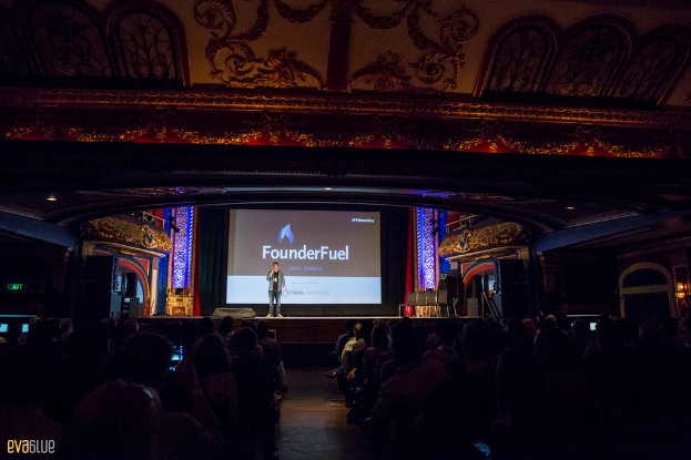 Eine Person steht auf einer Bühne mit einem Bildschirm im Hintergrund, schwarzen Vorhängen und einer Sitzreihe.