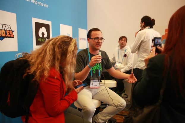 Ein Interview-Szenario mit Menschen mit Mikrofonen und einer Kamera, ein Banner links und Personen im Vordergrund und in der Mitte, mit einem klaren Hintergrund.