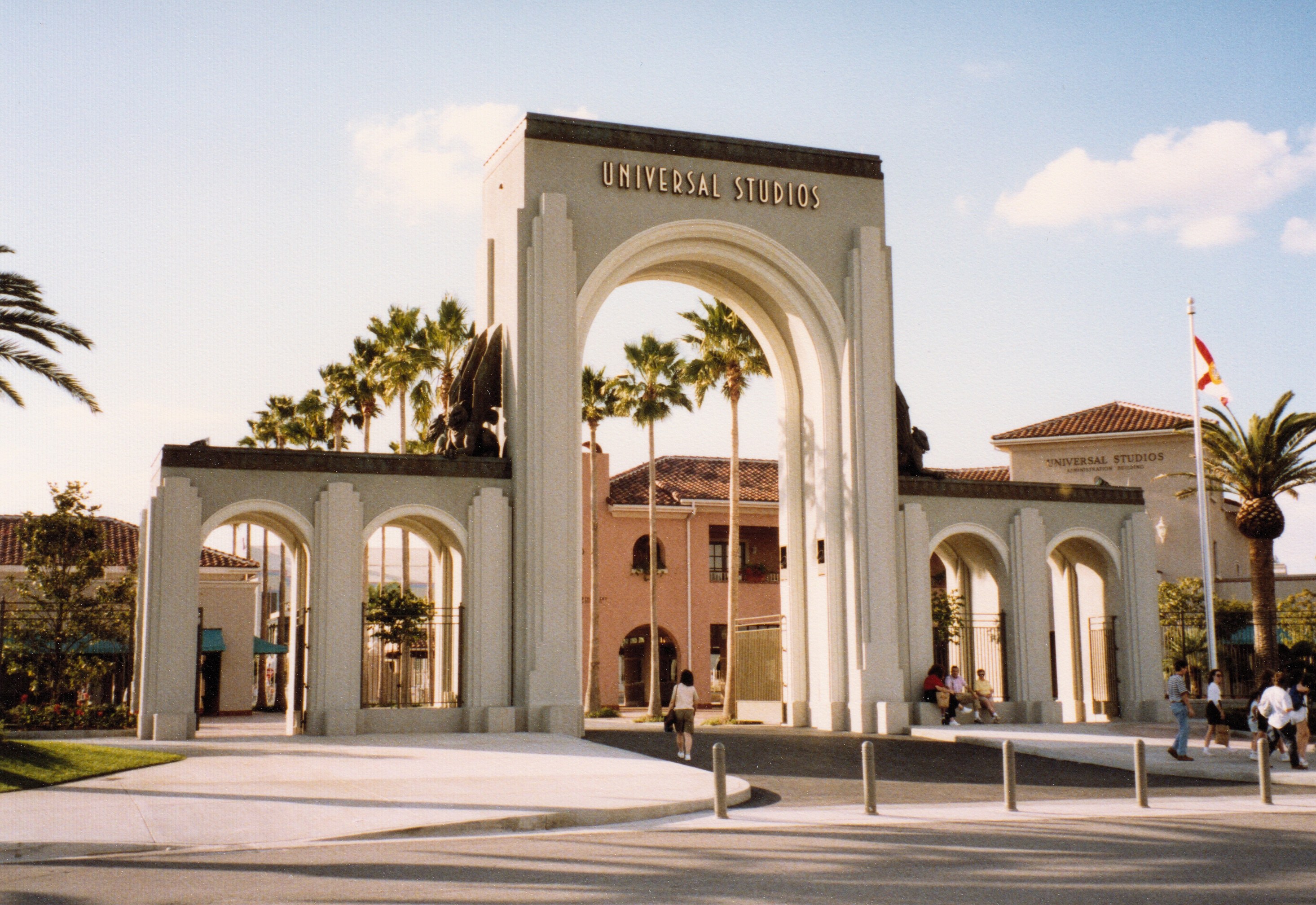 Eingang von Universal Studios Hollywood mit einem Bogen, einer Statue, Besuchern, Bäumen, Gebäuden, einem Fahnenmast und einem bewölkten Himmel.