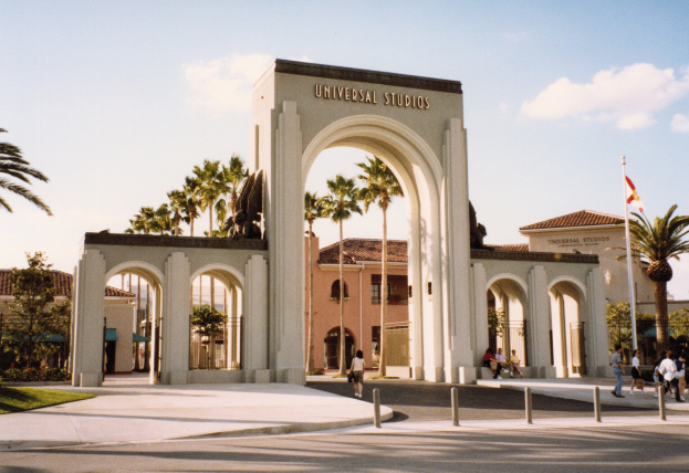 Eingang von Universal Studios Hollywood mit einem Bogen, einer Statue, Besuchern, Bäumen, Gebäuden, einem Fahnenmast und einem bewölkten Himmel.