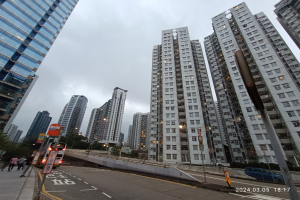 Eine Stadtstraße mit hohen Gebäuden, Fahrzeugen, Fußgängern, Schildern, Laternen, Bäumen und einem bewölkten Himmel, mit einem Wohngebäude im Vordergrund.