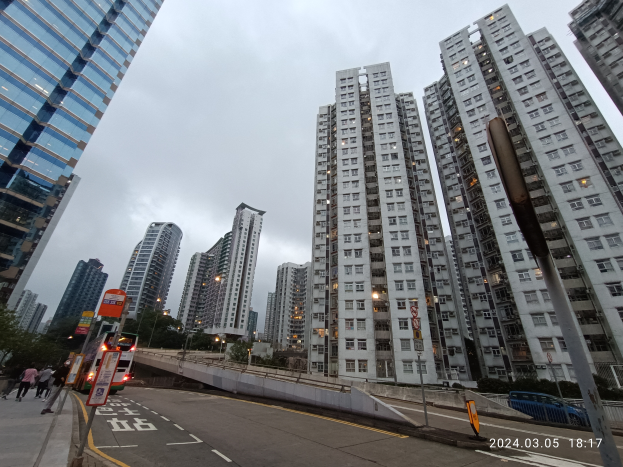 Eine Stadtstraße mit hohen Gebäuden, Fahrzeugen, Fußgängern, Schildern, Laternen, Bäumen und einem bewölkten Himmel, mit einem Wohngebäude im Vordergrund.