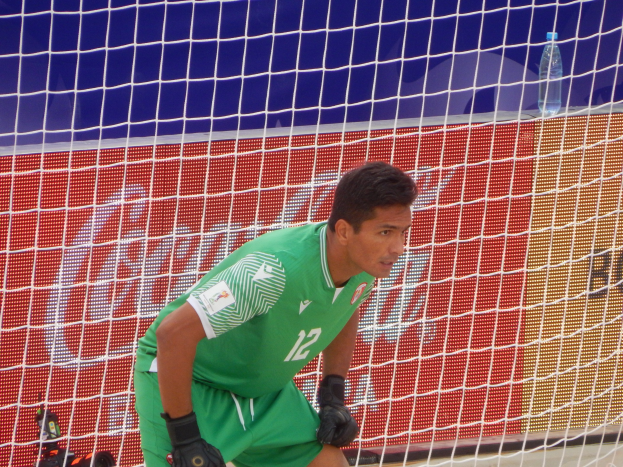 Ein Torwart in grüner Uniform kniet auf dem Boden neben einem Tor mit Netz im Hintergrund und einer Flasche rechts daneben.