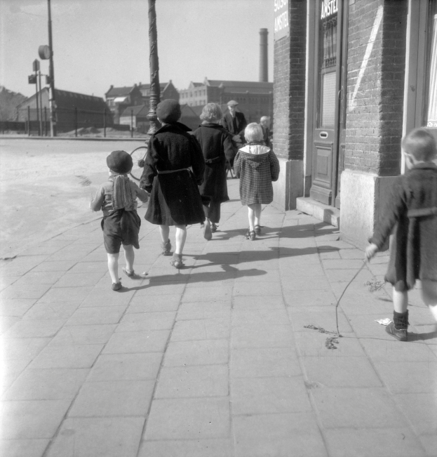 Eine Gruppe von Kindern, die auf einem Bürgersteig gehen, mit einem Gebäude auf der rechten Seite, das eine Tür und Fenster, Pfosten, Gebäude, Bäume und einen klaren Himmel im Hintergrund zeigt.