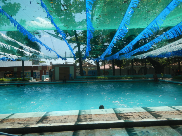 Großer Swimmingpool mit blauen und grünen Bändern an der Decke, Häuser und Bäume im Hintergrund, klarer blauer Himmel, Menschen schwimmen und genießen die Sonne.