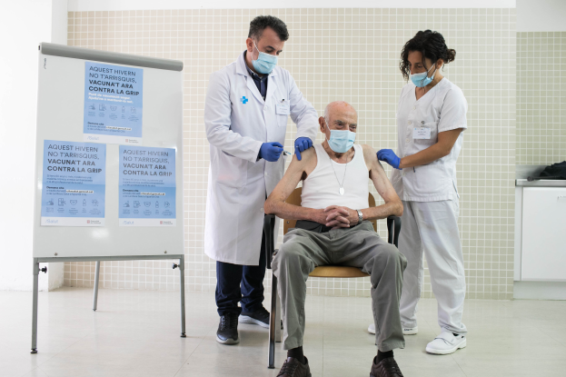 Ein älterer Mann sitzt in einem Stuhl und erhält von zwei maskierten und handschuhgetragenen Ärzten in einem Krankenhaus eine Impfung, mit einer Tafel links und Gegenständen auf einer Plattform rechts.