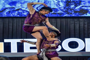 Zwei Performer in lila Kleidern führen einen akrobatischen Hebemanöver auf der Bühne vor einem Publikum aus