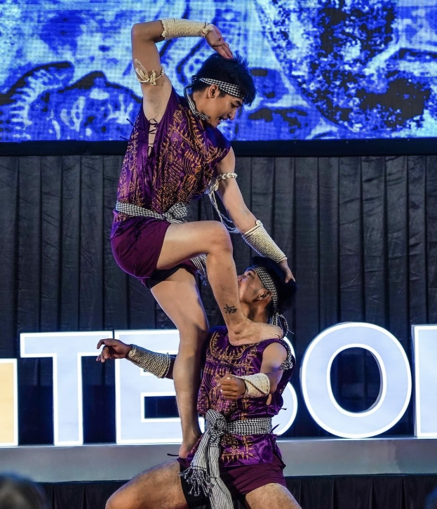 Zwei Performer in lila Kleidern führen einen akrobatischen Hebemanöver auf der Bühne vor einem Publikum aus