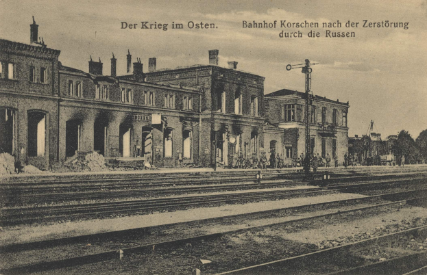 Schwarzes und weißes Foto des Bahnhofs Korschen in Ostern, Deutschland, das eine Bahnstation mit Gleisen, Gebäuden, Menschen, Bäumen und Himmel zeigt.