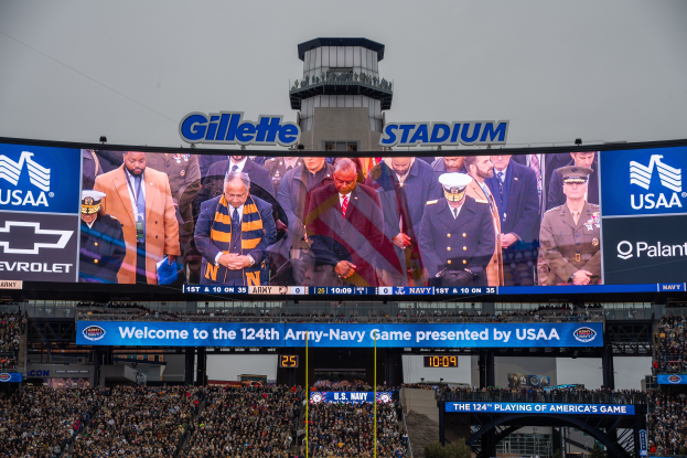 Eine große Menschenmenge in einem Stadion schaut ein Football-Spiel, mit einem zentralen Bildschirm, der Bilder und Text anzeigt, Schilder mit Text, Zuschauern in den Rängen, einem Bogen, Bäumen und einem klaren blauen Himmel im Hintergrund.