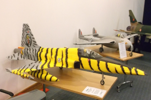 Verschiedene Flugzeugmodelle sind auf Tischen mit Etiketten angeordnet, ein Stuhl ist links und eine Wand im Hintergrund.