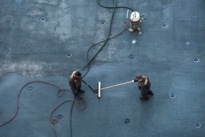 Zwei Männer auf einem Schiffdeck, jeder hält eine Pfeife in der Hand und verwendet eine Maschine, die über Kabel mit dem Deck verbunden ist, um die Deckoberfläche zu reinigen.