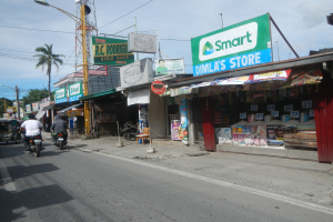 Eine belebte Straßenszene mit Motorradfahrern und Autos, umgeben von Gebäuden, Geschäften, Schildern, Strommasten, Kabeln, Straßenlaternen, Bäumen und einem bewölkten Himmel.